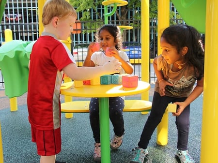 three small children one boy and two girls sitting at a yellow tables playing with blocks outside at a park-1