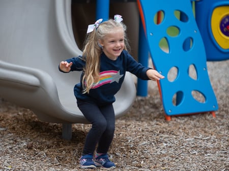 young girl in a navy sweater with a star on it with two white bow ties in her hair smiling getting off of a gray slide at park